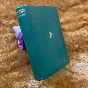 May include: A green hardback book with the title "The Gap in the Curtain" by John Buchan. The book has gold lettering and a gold "JB" monogram on the front cover.