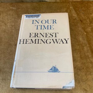 May include: A hardcover book titled In Our Time by Ernest Hemingway with a cream-colored dust jacket. The cover features blue text, a small blue sailboat illustration, and a library barcode sticker at the top with the number 3 4028 03310 1628.