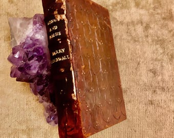 May include: An antique book titled "SONG AND POEMS" by Barry Cornwall, with gold lettering on a dark spine. The cover is brown with a textured pattern. The book rests against a cluster of purple amethyst crystals.