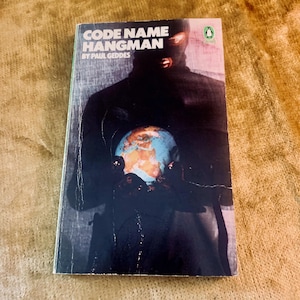 May include: A paperback book titled "CODE NAME HANGMAN" by Paul Geddes. The cover shows a masked figure holding a globe. The Penguin Books logo is in the top right corner. The book has a vintage aesthetic.