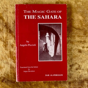 Pode incluir: Uma capa de livro vermelha com o título "O Portal Mágico do Saara" de Angelo Piccioli. A capa do livro apresenta uma fotografia em preto e branco de uma rua estreita numa cidade com um alto edifício branco ao longe.