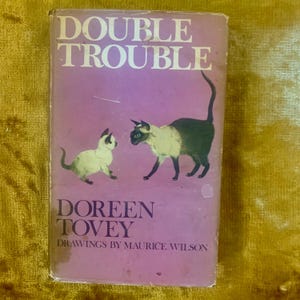 Op de afbeelding: Een roze boekomslag met de titel "Double Trouble" in witte letters. De omslag toont een illustratie van twee Siamese katten, een groter dan de andere, die elkaar aankijken. De naam van de auteur, "Doreen Tovey", is in zwart onder de illustratie gedrukt. De tekst "Tekeningen van Maurice Wilson" is in kleinere zwarte letters onder de naam van de auteur gedrukt.