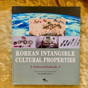 May include: A green book titled "Korean Intangible Cultural Properties: Traditional Handicrafts" by the Cultural Properties Administration, The Republic of Korea. The book cover features a collage of images including a traditional Korean painting, a woman weaving, and a collection of traditional Korean crafts.