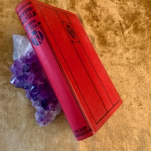 May include: A vintage book with a red cover, titled "Greenmantle" by John Buchan, rests against a cluster of purple amethyst crystals. The book's spine displays the title and author's name in black lettering. The background is a textured gold surface.