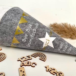 Könnte beinhalten: Ein grauer Filzhut mit goldenen Sternen und Flaggen. Der Hut hat den Text "Bye Bye Grundschule" und "Hallo 1. Klasse 2024" aufgedruckt. Der Hut ist umgeben von Holz-Ausschnitten von Zahlen, Buchstaben und Regenbögen.