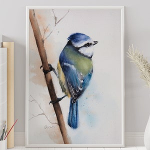 May include: Watercolour painting of a blue tit bird on a branch. The bird features blue, yellow, and green plumage. The artwork is framed in white and set against a neutral backdrop. A white vase with dried flowers is to the right.
