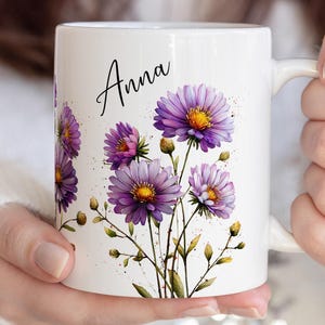 May include: White ceramic mug featuring a floral design with purple and yellow flowers and green stems. The name "Anna" is written in black script. The mug is held by a person.