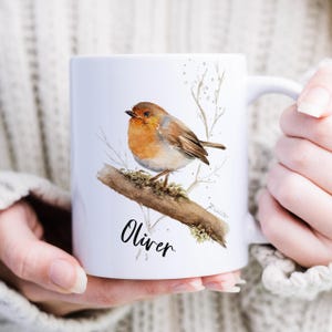 Puede incluir: Taza de cerámica blanca con una ilustración de acuarela de un petirrojo posado en una rama. El pájaro tiene plumas naranjas y marrones. El nombre "Oliver" está escrito en cursiva debajo. La taza es sostenida por una persona.