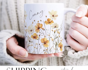 Könnte beinhalten: Eine weiße Keramiktasse mit einem Blumendesign mit gelben und weißen Blumen und grünen Stielen. Die Tasse trägt den Namen "Conny" in schwarzer Kursivschrift.