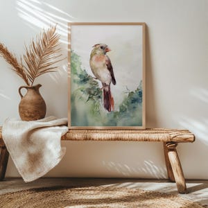 Könnte beinhalten: Aquarellmalerei eines Vogels auf einem belaubten Ast, gerahmt in hellem Holz. Der Vogel hat einen beigen Körper, braune Flügel und einen roten Schwanz. Der Hintergrund ist ein weiches Grün und Weiß. Das Kunstwerk wird auf einer gewebten Bank ausgestellt.