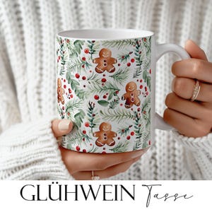 May include: White ceramic mug with a festive gingerbread man and botanical pattern. The design features brown gingerbread men, green leaves, and red berries on a white background. The mug is held by a person wearing a white sweater. The text "GLÜHWEIN Tasse" is at the bottom.