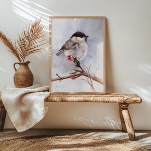 Könnte beinhalten: Ein gerahmtes Aquarellgemälde eines kleinen Vogels, der auf einem Ast mit rosa Blüten sitzt. Der Vogel hat einen schwarzen Kopf und einen grauen und weißen Körper. Das Kunstwerk wird auf einer gewebten Bank mit einer Vase und einer getrockneten Pflanze ausgestellt.