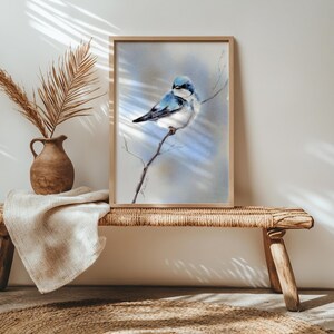 May include: A framed watercolour print of a blue and white bird perched on a branch. The artwork has a soft, painterly style with a light blue and grey background. The frame is a light wood colour. The print is displayed on a woven bench.