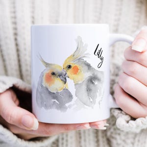 Könnte beinhalten: Weiße Keramik-Tasse mit einer Aquarell-Illustration von zwei sich küssenden Nymphensittichen. Die Vögel haben gelbe Hauben, orangefarbene Wangenflecken und graue Körper. Der Name "Lilly" ist in schwarzer Schrift auf der Tasse geschrieben.