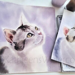 May include: A watercolor painting of a grey and white tabby cat with yellow eyes looking upward against a soft purple background. The artwork features the text @KathleensWatercolor. A smaller print of the same cat portrait is positioned to the right.