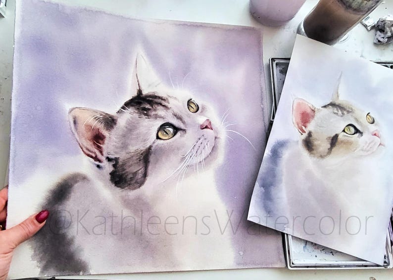 May include: A watercolor painting of a grey and white tabby cat with yellow eyes looking upward against a soft purple background. The artwork features the text @KathleensWatercolor. A smaller print of the same cat portrait is positioned to the right.