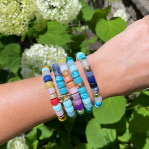Puede incluir: Un conjunto de pulseras de cuentas de colores con una variedad de piedras y cuentas. Las pulseras est&aacute;n superpuestas en la mu&ntilde;eca de una persona y presentan cuentas turquesa, moradas, rosas, amarillas y azules.