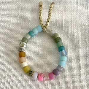 K&ouml;nnte beinhalten: Ein farbenfrohes Perlenarmband mit einem goldenen Kordelverschluss. Die Perlen sind in verschiedenen Blaut&ouml;nen, Gr&uuml;nt&ouml;nen, Gelb, Rosa und Lila.