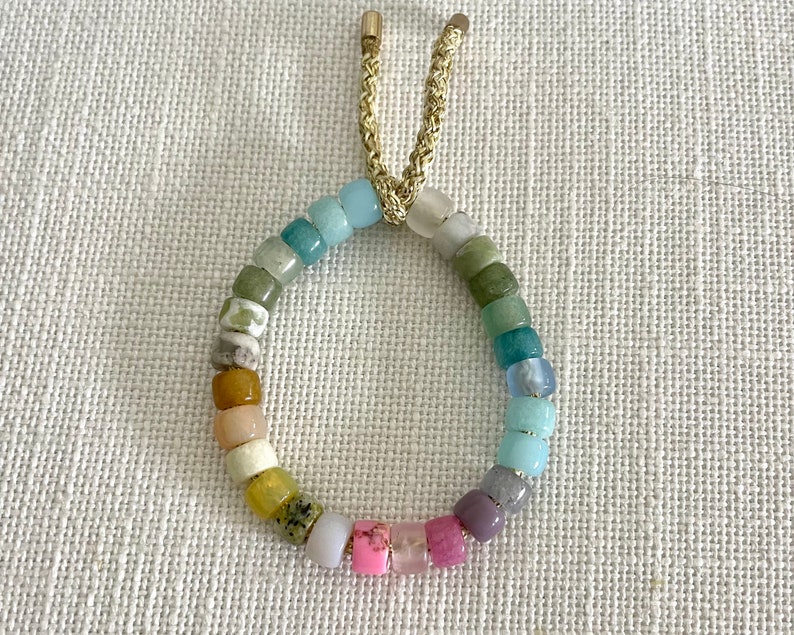 K&ouml;nnte beinhalten: Ein farbenfrohes Perlenarmband mit einem goldenen Kordelverschluss. Die Perlen sind in verschiedenen Blaut&ouml;nen, Gr&uuml;nt&ouml;nen, Gelb, Rosa und Lila.