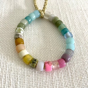 K&ouml;nnte beinhalten: Ein farbenfrohes Perlenarmband mit einem Regenbogen an Farben, darunter Rosa, Lila, Gr&uuml;n, Blau und Gelb. Das Armband ist aus nat&uuml;rlichen Steinen gefertigt und hat einen goldenen Kordelverschluss.