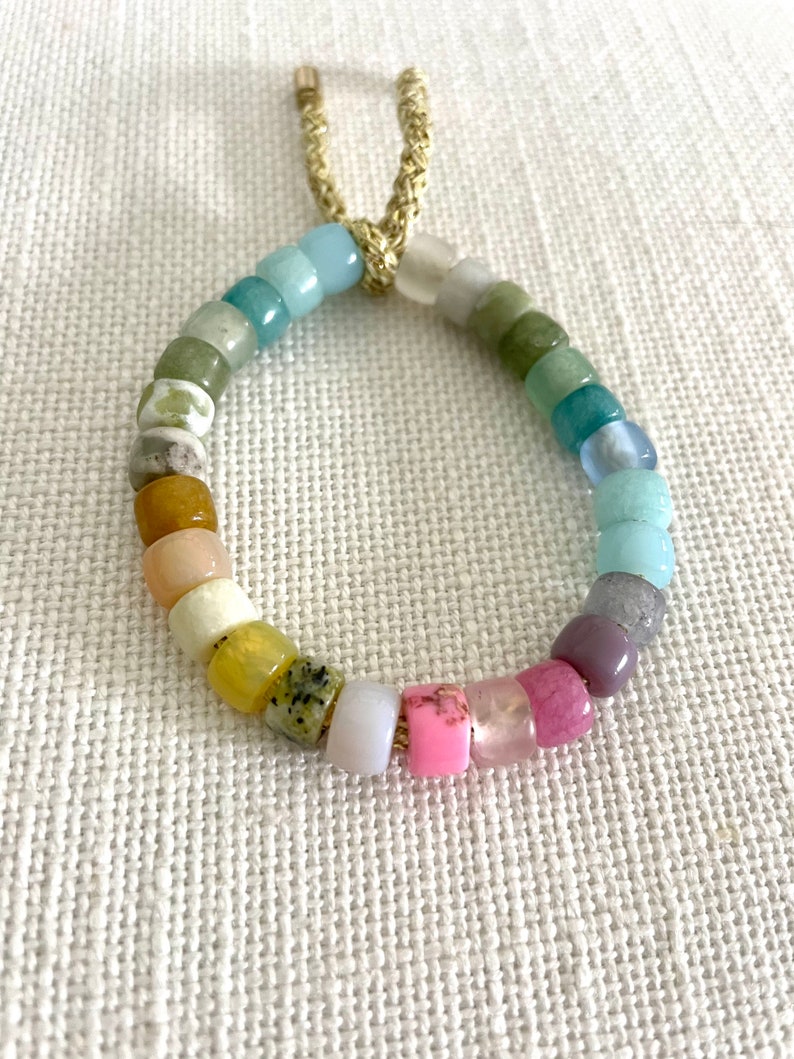 K&ouml;nnte beinhalten: Ein farbenfrohes Perlenarmband mit einem Regenbogen an Farben, darunter Rosa, Lila, Gr&uuml;n, Blau und Gelb. Das Armband ist aus nat&uuml;rlichen Steinen gefertigt und hat einen goldenen Kordelverschluss.