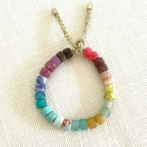 May include: A rainbow colored beaded bracelet with a gold cord closure. The beads are square shaped and are arranged in a rainbow pattern.