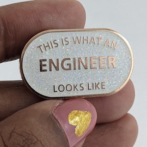 May include: A white enamel pin with rose gold lettering that reads "This is what an engineer looks like". The pin has a glittery finish.