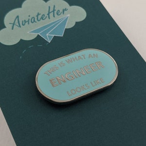 May include: A teal blue card with a white cloud and a paper aeroplane. The card says "AviateHer" and has a blue enamel pin that says "This is what an Engineer looks like".