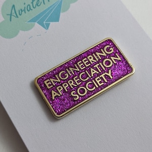 Op de afbeelding: Een gouden pin met een paarse glitterachtergrond. De pin heeft de tekst "Engineering Appreciation Society".