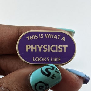 Puede incluir: Un pin ovalado de esmalte morado y dorado con el texto "THIS IS WHAT A PHYSICIST LOOKS LIKE". El pin está sujeto por una mano con esmalte de uñas azul y turquesa.