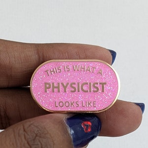 Puede incluir: Un pin de esmalte rosa brillante con ribete dorado. El pin dice "THIS IS WHAT A PHYSICIST LOOKS LIKE" en letras doradas. El pin tiene forma ovalada.