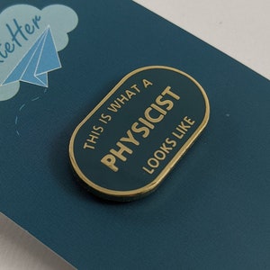 Puede incluir: Un pin dorado con el texto "THIS IS WHAT A PHYSICIST looks like" en una tarjeta verde azulado con un logotipo de nube y avión de papel. El pin tiene forma ovalada con un fondo azul oscuro y letras doradas.