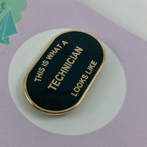 Może przedstawiać: Złota i czarna emaliowana przypinka z tekstem "This is what a technician looks like".
