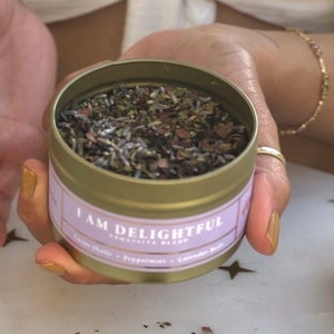 May include: A gold metal tin with a purple label that reads "I AM DELIGHTFUL" and a list of ingredients. The tin is filled with dried herbs and flowers.