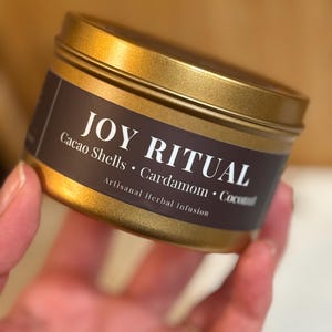 May include: A gold-colored tin with a dark brown label. The label reads "JOY RITUAL" in white text, with the ingredients "Cacao Shells • Cardamom • Coconut" listed below. The words "Artisanal Herbal Infusion" are also visible.