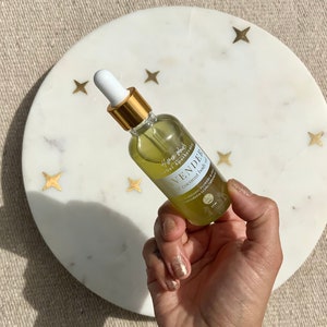 Op de afbeelding: Een glazen flesje met lavendel-kokosolie voor het lichaam met een gouden dop en druppelaar. De fles is gelabeld met "Here & Now Apothecary" en "Lavender Coconut Body Oil".