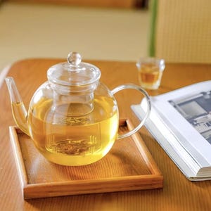以下が含まれることがあります： 金色の紅茶が入った透明なガラスのティーポット。内蔵のインフューザーと丸みを帯びたハンドルが付いています。ティーポットは木製のトレイの上にあり、近くには小さなグラスの紅茶と本があります。シーンは木製のテーブルに設定されています。