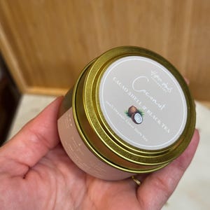 Könnte beinhalten: Eine runde Dose mit goldenem Rand mit Kokosnuss-Kakaoschalen- und Schwarztee. Das Etikett zeigt das Wort "Coconut" in einer Schreibschrift, zusammen mit einer Illustration einer Kokosnuss. Die Dose wird in einer Hand gehalten.