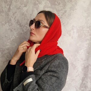May include: A woman wearing a dark gray wool coat and a bright red scarf. The coat has a classic design with large buttons and pockets. She is also wearing sunglasses and a watch. The background is a textured gray wall.
