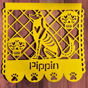 May include: A bright yellow rectangular paper cutout featuring a central skeleton dog silhouette, two decorative skulls, and the name Pippin cut out in the center. The border has a scalloped edge with four paw print cutouts along the bottom.