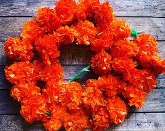 Puede incluir: Una guirnalda circular construida con numerosas flores de caléndula artificiales de color naranja vibrante. Las flores individuales están densamente agrupadas y presentan pétalos rizados y superpuestos. Un tubo de plástico verde une las flores.
