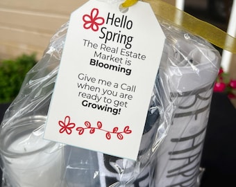 May include: A gift bag with a white tag that reads "Hello Spring. The Real Estate Market is Blooming. Give me a Call when you are ready to get Growing!" The bag contains a white candle, a white towel with black text that reads "Be Happy", and a black and white patterned tote bag.