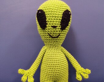 May include: A green crocheted alien toy with black eyes and a smiling mouth. The toy is standing on a blue surface.