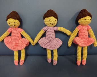 May include: Three crocheted dolls wearing pink and purple dresses. The dolls have brown hair and yellow faces with black eyes and smiles.