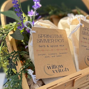 May include: Two white muslin bags filled with herbs and spices, labeled "Springtime Simmer Pot" with a list of ingredients. The bags are in a woven wicker basket with lavender sprigs and green foliage.