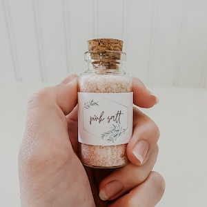 Peut inclure: Un petit pot en verre avec un bouchon en liège rempli de sel rose de l'Himalaya. Le pot a une étiquette blanche avec des feuilles vertes et le texte "pink salt".