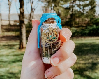 May include: A small glass bottle with a blue wax seal and a gold charm featuring a lotus flower design. The bottle is filled with various dried herbs and flowers.