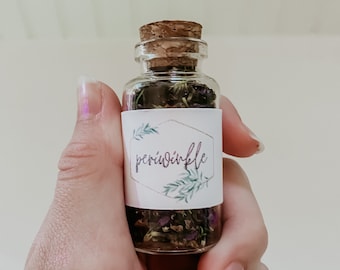 May include: A small glass bottle with a cork stopper filled with dried herbs and flowers. The bottle has a white label with the word "periwinkle" written in black.