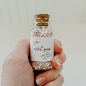 May include: A small glass bottle with a cork top filled with white eggshell powder. The bottle has a white label with green leaves and the text "eggshell powder".