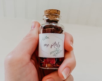 May include: A small glass bottle with a cork stopper filled with dried rose petals. The bottle has a white label with the text "rose petals" in black lettering.